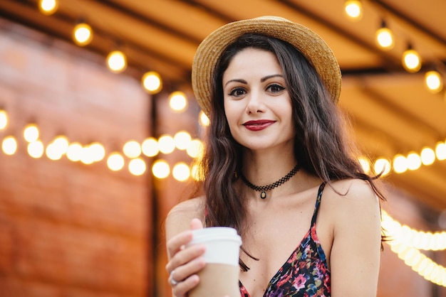 Ritratto di donna bruna con occhi affascinanti labbra rosse ben modellate e pelle pura vestita con cappello estivo e camicia con caffè da asporto fresco in tazza di carta rilassante sulla terrazza dell'accogliente caffè