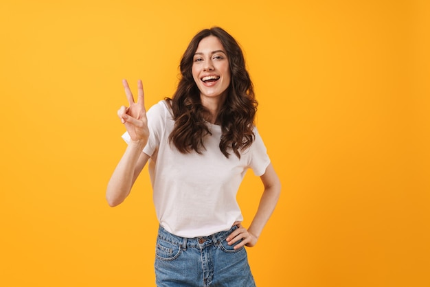 Ritratto di donna bruna felice che indossa abiti casual sorridendo e mostrando segno di pace in telecamera isolata su muro giallo