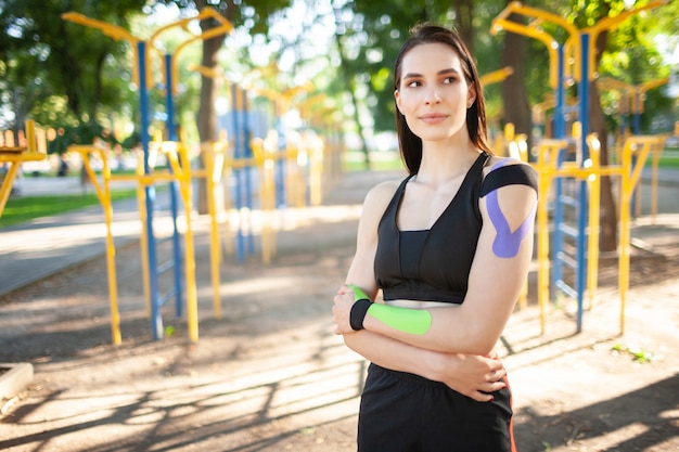 Ritratto di donna bruna muscolare attraente che indossa abiti sportivi neri, guardando la fotocamera. Giovane atleta femminile sorridente in posa con le braccia incrociate, kinesiotaping colorato sul corpo.