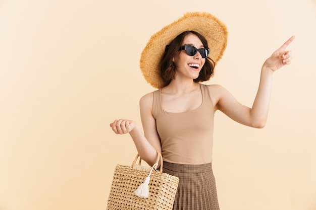 Ritratto di donna bruna sorridente che indossa cappello di paglia e occhiali da sole in posa con borsa estiva mentre punta il dito contro copyspace isolato