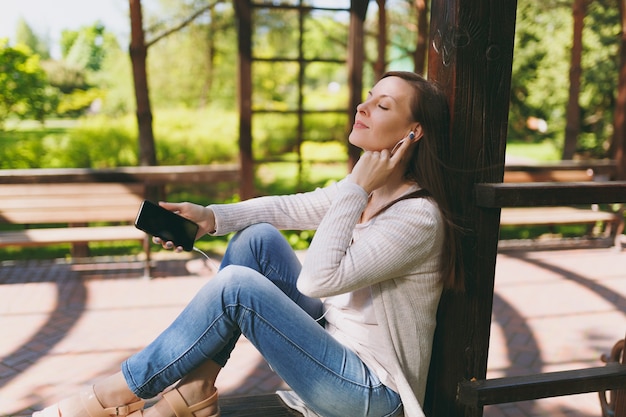 Ritratto di donna calma in abiti casual. Donna seduta nel parco in strada all'aperto sulla natura, ascoltando musica nel telefono cellulare con schermo vuoto vuoto per copiare lo spazio negli auricolari. Concetto di stile di vita.