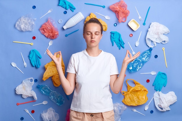 Ritratto di donna calma vestita di maglietta bianca ha raccolto rifiuti di plastica isolati sulla parete blu praticando yoga cercando di calmare le preoccupazioni per l'ambiente e causando danni alla natura