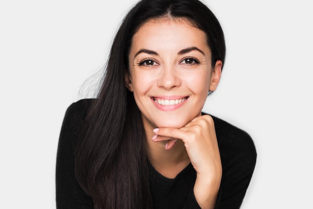 Ritratto di donna carina bruna con un sorriso a trentadue denti bello e sano con la mano sul mento Giovane donna graziosa felice che indossa un maglione nero su sfondo grigio chiaro Umore positivo e di felicità