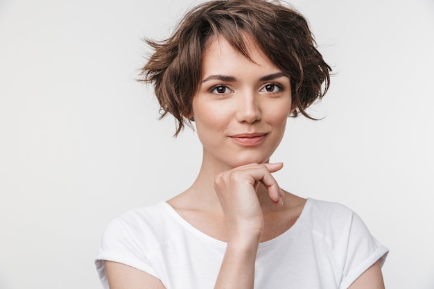 Ritratto di donna carina con capelli corti castani in maglietta di base mentre si trova isolata su un muro bianco