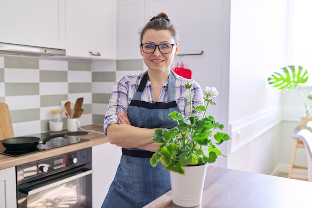 Ritratto di donna casalinga con fiori in vaso in cucina a casa. Femmina matura in grembiule che guarda l'obbiettivo