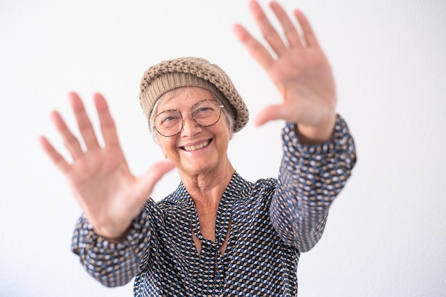 Ritratto di donna caucasica anziana che indossa cappello e occhiali isolati su sfondo bianco facendo il gesto di arresto della mano