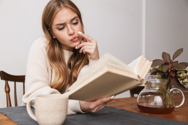 Ritratto di donna caucasica confusa che indossa abiti casual leggendo un libro e bevendo tè in una stanza accogliente