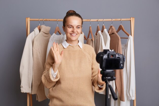 Ritratto di donna caucasica felice sorridente in piedi vicino a appendiabiti con abiti nel negozio di moda che registra video per il suo blog che riprende con la fotocamera sul treppiede agitando la mano che dice ciao