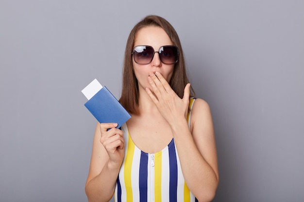 Ritratto di donna caucasica stupita che indossa costume da bagno e occhiali da sole in possesso di passaporto con biglietti sorpresi coprendo la bocca con la mano isolata su sfondo grigio ha vacanze all'estero