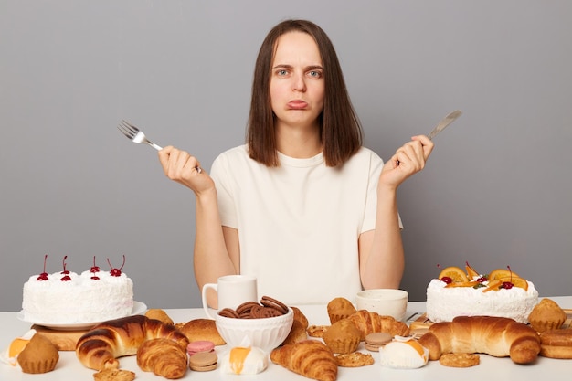 Ritratto di donna caucasica triste stressata con i capelli castani seduta al tavolo isolata su sfondo grigio che guarda l'obbiettivo con le labbra imbronciate sconvolte mantiene la dieta vuole panetteria dolce