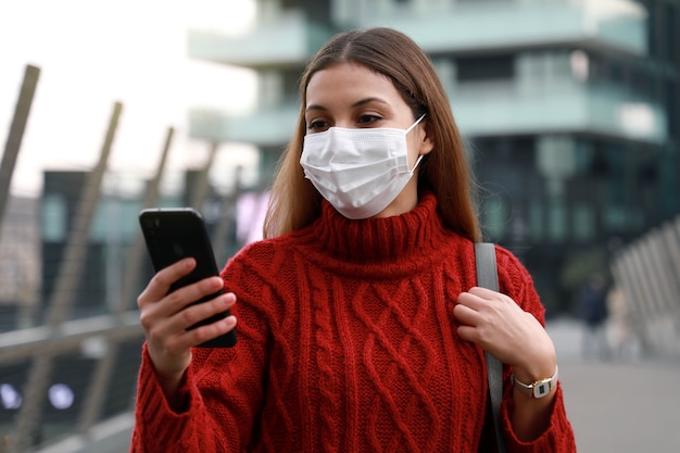 Ritratto di donna con maschera protettiva a piedi nella città moderna utilizzando smartphone