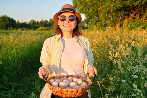 Ritratto di donna contadina in cappello con cesto di uova fresche. Agricoltura, allevamento di prodotti ecologici biologici. Strada di campagna, campagna, tramonto, natura, sfondo del giardino