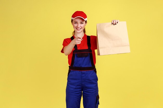 Ritratto di donna corriere che tiene pacco ordinato consegna cibo in sacchetto di carta che punta il dito verso la fotocamera indossando tute e cappuccio Studio indoor girato isolato su sfondo giallo