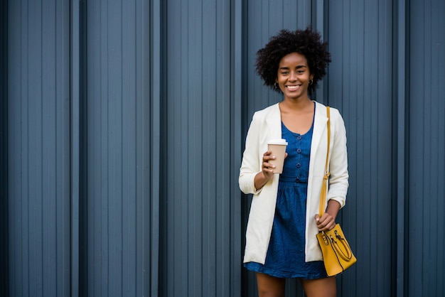 Ritratto di donna d'affari afro che tiene una tazza di caffè mentre levandosi in piedi all'aperto sulla strada. Business e concetto urbano.