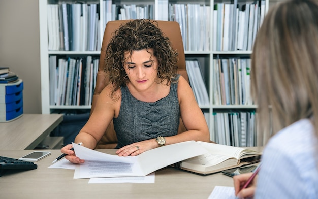 Ritratto di donna d'affari riccia che lavora con documenti in ufficio mentre segretaria bionda prende appunti taking