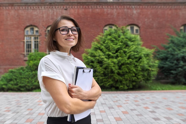 Ritratto di donna d'affari sorridente sicura, insegnante, consulente con documenti cartacei appunti. Donna sorridente positiva, spazio per la copia, edificio scolastico, sfondo dell'edificio per uffici