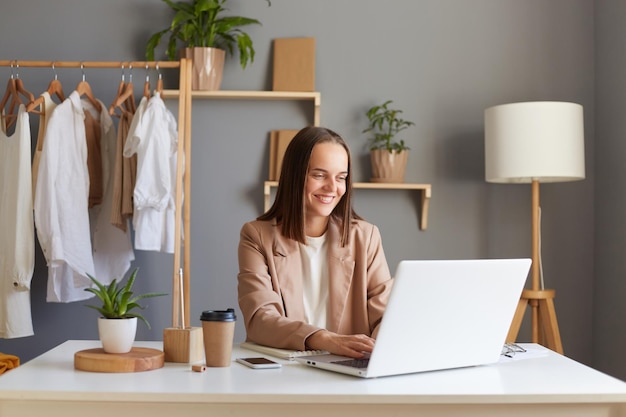 Ritratto di donna dai capelli castani positiva sorridente che indossa una giacca beige seduta al suo posto di lavoro davanti al computer portatile che digita sulla tastiera guardando il display