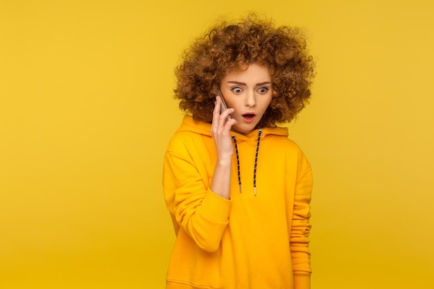 Ritratto di donna dai capelli ricci sorpresa in felpa con cappuccio in stile urbano che chiama un amico che parla sullo smartphone con espressione stupita che discute di notizie scioccanti in studio al coperto isolato su sfondo giallo