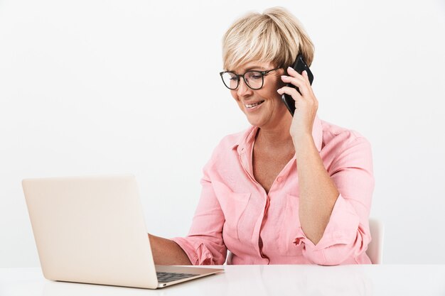 Ritratto di donna di mezza età caucasica che indossa occhiali da vista che parla al cellulare e utilizza un computer portatile isolato su un muro bianco