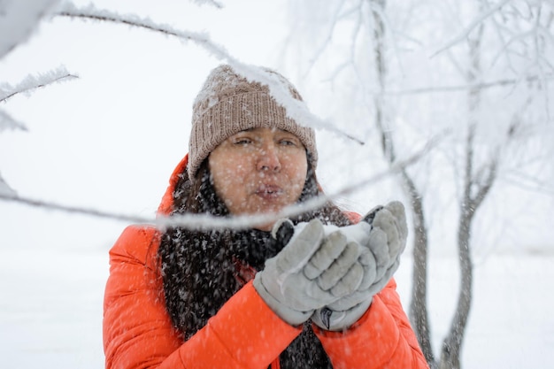 Ritratto di donna di mezza età che soffia via i fiocchi di neve guanti con ramoscelli sfocati ricoperti di neve in primo piano sulla passeggiata nella foresta innevata Magica ora invernale piena di colore bianco