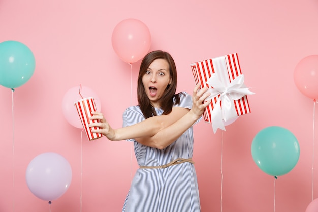 Ritratto di donna divertente in abito blu che tiene scatola rossa con regalo regalo e tazza di plastica di soda o cola su sfondo rosa con mongolfiere colorate. Festa di compleanno, persone sincere emozioni.