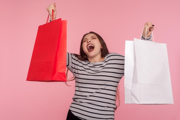 Ritratto di donna entusiasta euforica ed entusiasta in felpa a righe che urla, esprime grande gioia e solleva borse della spesa, deliziata dalle vendite nel negozio di moda. girato in studio al coperto, isolato