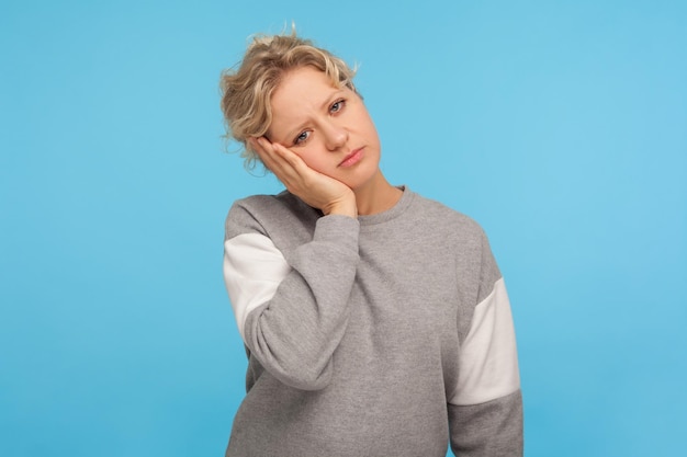 Ritratto di donna esausta annoiata con capelli corti ricci in felpa appoggiata a una mano e guardando la fotocamera con disinteresse stanco di una noiosa storia triste in studio indoor girato isolato su sfondo blu