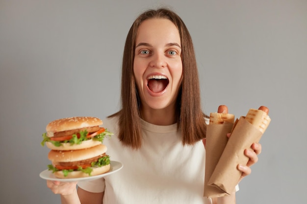 Ritratto di donna estremamente felice che mangia fast food tenendo hot dog e sandwich indossando una maglietta bianca in posa isolata su sfondo grigio esultando per avere un pasto cheat