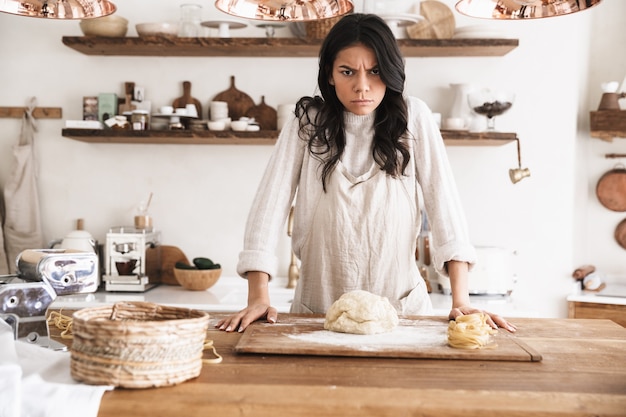 Ritratto di donna europea irritata che indossa un grembiule che cucina e prepara pasta fatta in casa in cucina a casa