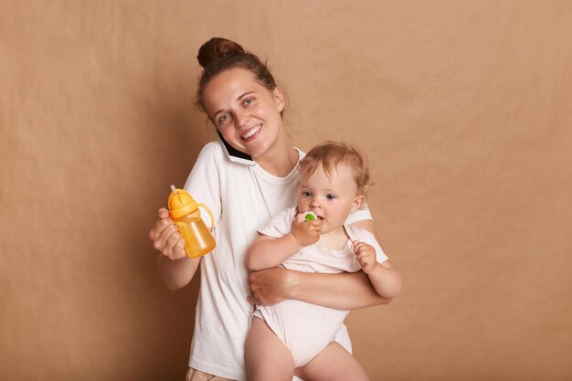 Ritratto di donna felice attraente che indossa abbigliamento casual in piedi con il suo bambino bambino isolato su sfondo marrone guardando la fotocamera e parlando di smart phone