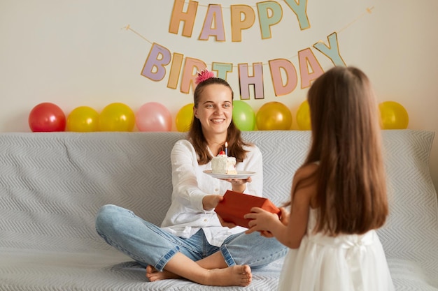 Ritratto di donna felice che celebra il suo compleanno piccola figlia carina che posa all'indietro alla macchina fotografica e che dà alla madre il suo regalo nella decorazione festiva della scatola rossa sullo sfondo