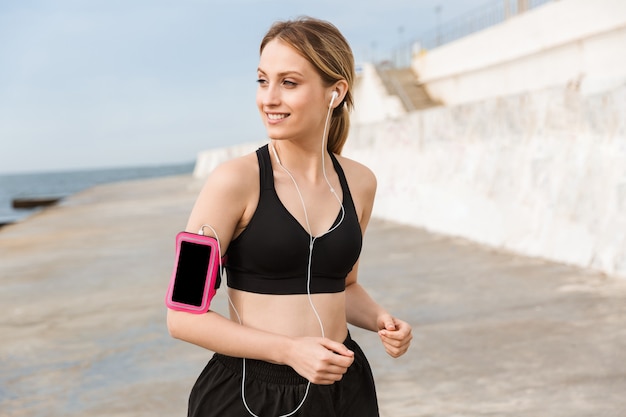 Ritratto di donna felice che indossa una custodia per smartphone che ascolta musica con gli auricolari mentre fa jogging vicino al mare al mattino