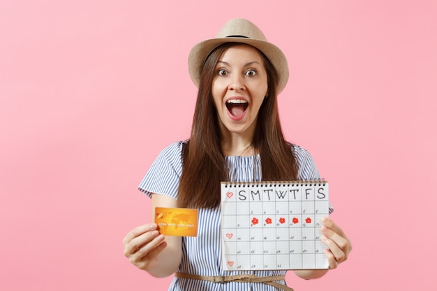 Ritratto di donna felice in abito blu, cappello con carta di credito, calendario dei periodi, controllo dei giorni delle mestruazioni isolati su sfondo rosa di tendenza. Concetto ginecologico sanitario medico. Copia spazio.