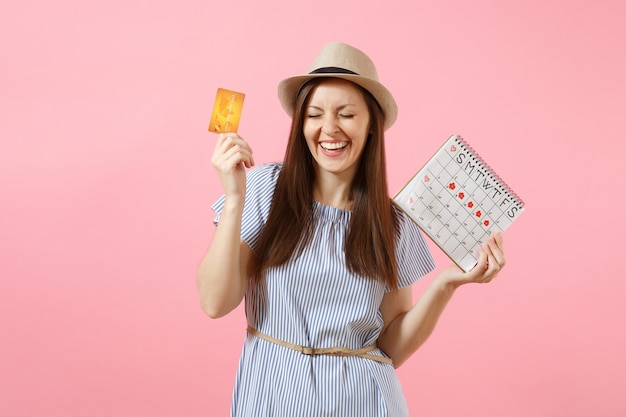 Ritratto di donna felice in abito blu, cappello con carta di credito, calendario dei periodi, controllo dei giorni delle mestruazioni isolati su sfondo rosa di tendenza. Concetto ginecologico sanitario medico. Copia spazio.