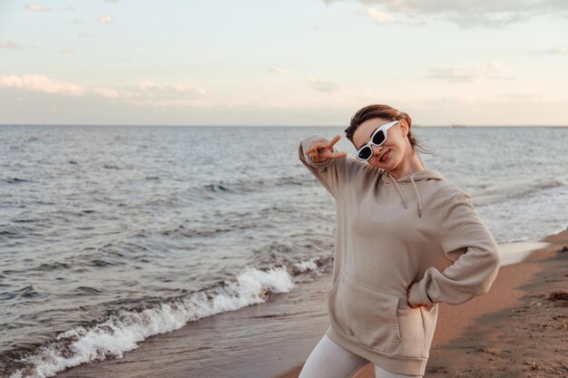 Ritratto di donna felice in felpa con cappuccio e occhiali da sole sulla spiaggia che mostra il segno di pace