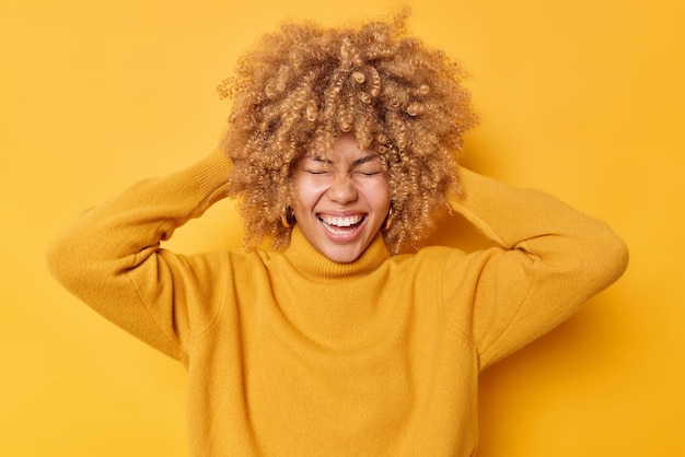 Ritratto di donna felicissima con capelli folti biondi tiene le mani sulla testa ride con gioia indossa un maglione casual tiene gli occhi chiusi isolati su sfondo giallo. Scatto monocromatico. Concetto di emozioni