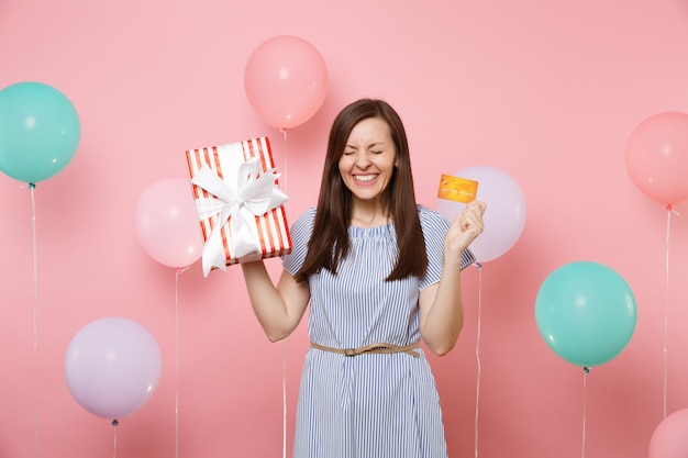 Ritratto di donna felicissima con gli occhi chiusi in abito blu in possesso di carta di credito e scatola rossa con regalo presente su sfondo rosa con mongolfiera colorata. Festa di compleanno, persone sincere emozioni.