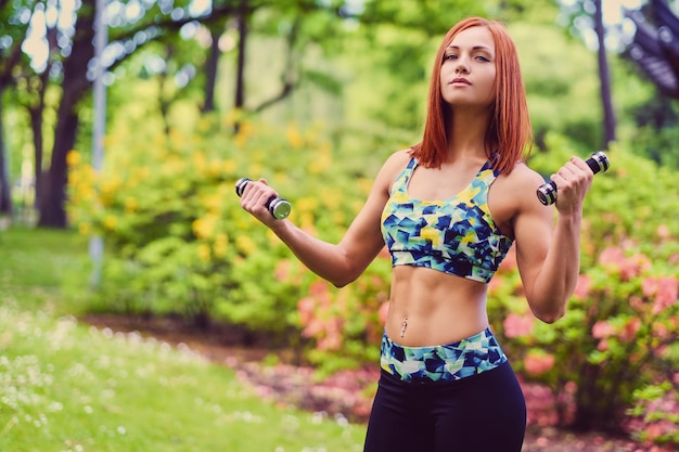 Ritratto di donna fitness rossa tiene manubri in un parco all'aperto.