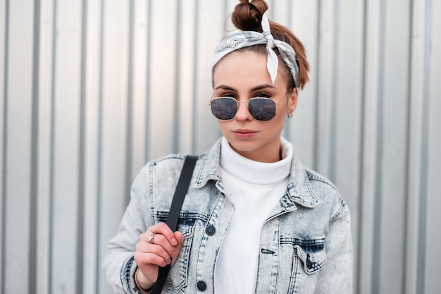 Ritratto di donna giovane hipster in occhiali da sole alla moda con un'acconciatura alla moda in un maglione vintage bianco lavorato a maglia in una giacca di jeans con uno zaino nero vicino a un muro di metallo. Ragazza moderna americana.