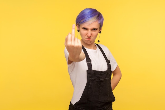 Ritratto di donna hipster arrabbiata pazza con capelli viola in tuta di jeans che mostra il dito medio maleducato scopare il viso pieno di odio e aggressività isolato su sfondo giallo girato in studio