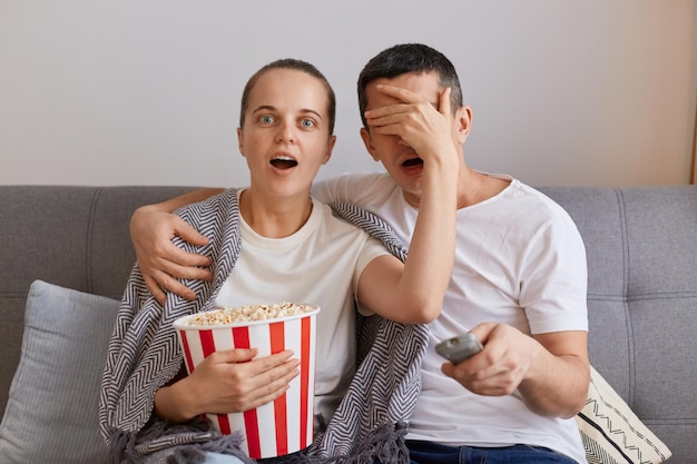 Ritratto di donna impressionata guardando film horror con uomo scioccato moglie coperta occhi mariti con palmo proibito contenuto in tv coppia seduta sul divano avvolta in una coperta