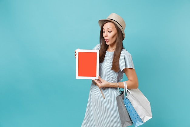 Ritratto di donna in abito estivo, cappello di paglia che tiene pacchetti borse con acquisti dopo lo shopping, computer tablet pc con schermo vuoto isolato su sfondo blu pastello. Copia spazio per la pubblicità.