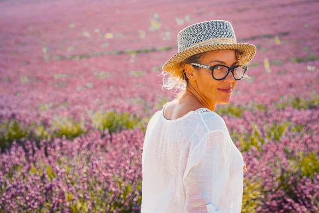 Ritratto di donna in cappello in piedi sul campo di fiori di lavanda. Bella donna in occhiali sorridente mentre si sta in piedi sul campo. Donna che osserva sopra la spalla dal campo di fiori di lavanda agricolo