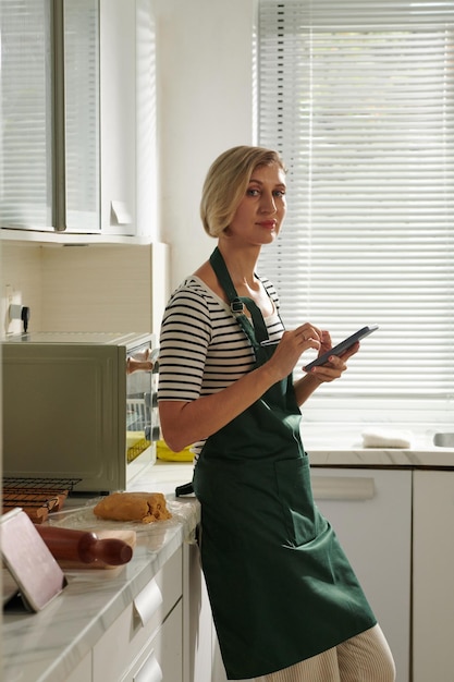 Ritratto di donna in grembiule verde che impara sul bancone della cucina durante la lettura della ricetta sul tablet PC