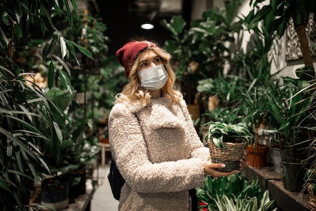 Ritratto di donna in maschera medica protettiva, nuovo normale shopping al dettaglio, scelta per l'acquisto di vaso di fiori con felce per regalo in negozio di fiori. Distanza sociale, stile di vita. Nuovo concetto normale