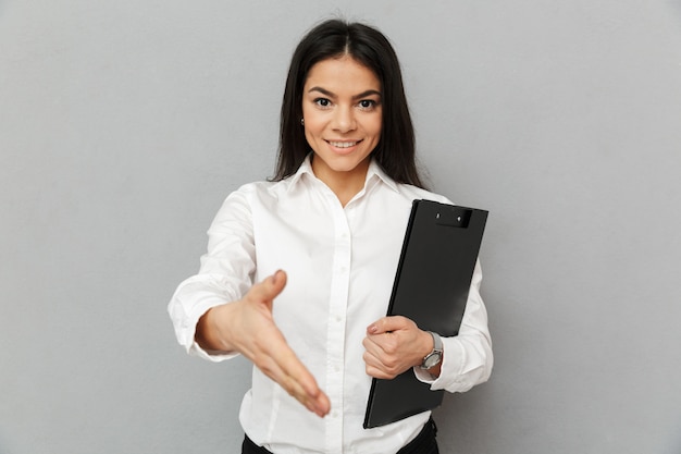 Ritratto di donna in ufficio con lunghi capelli scuri che indossa una camicia bianca sorridente e dando la mano per tremare mentre si tiene la cartella con i documenti, isolate su uno sfondo grigio