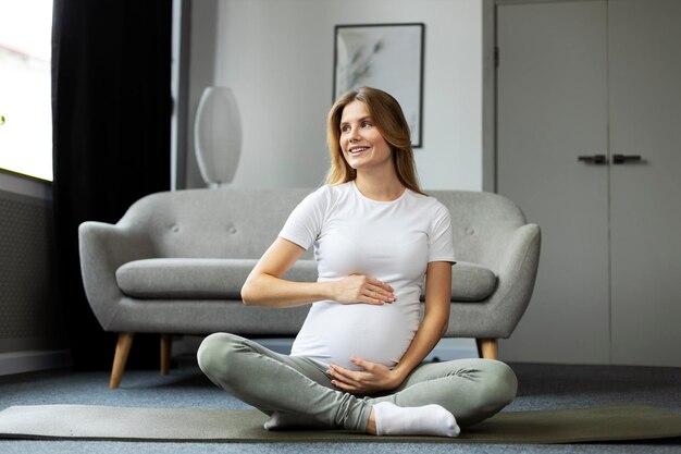 Ritratto di donna incinta sorridente seduta sul pavimento e toccante al ventre Gravidanza yoga