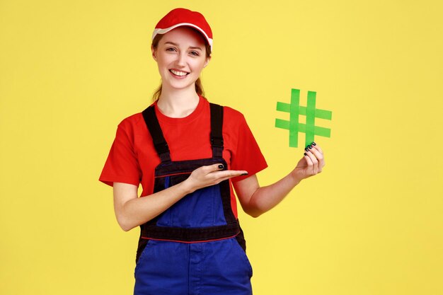 Ritratto di donna lavoratrice in piedi che presenta hashtag verde nelle mani guardando la fotocamera con espressione positiva che indossa tute e berretto rosso Studio indoor girato isolato su sfondo giallo