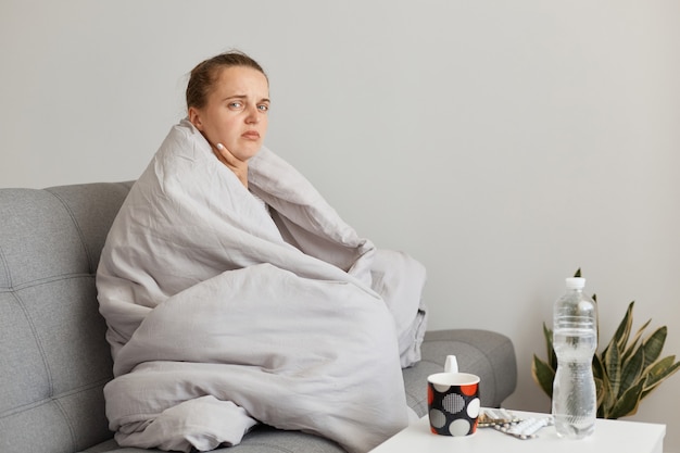 Ritratto di donna malata con chignon seduto sul divano avvolto in una coperta che guarda l'obbiettivo con espressione facciale triste, toccando il suo dolore al collo, soffrendo di mal di gola.