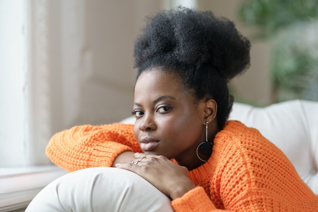 Ritratto di donna millenaria afroamericana alla moda in abbigliamento casual e capelli ricci sdraiato sul divano, rilassante a casa. Studentessa etnica abbastanza premurosa in maglione arancione che guarda l'obbiettivo.